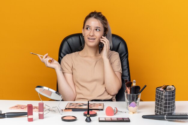 Smiling looking side young beautiful girl wearing dental braces sits at table with makeup tools holding makeup brush speaks on phone isolated on orange background