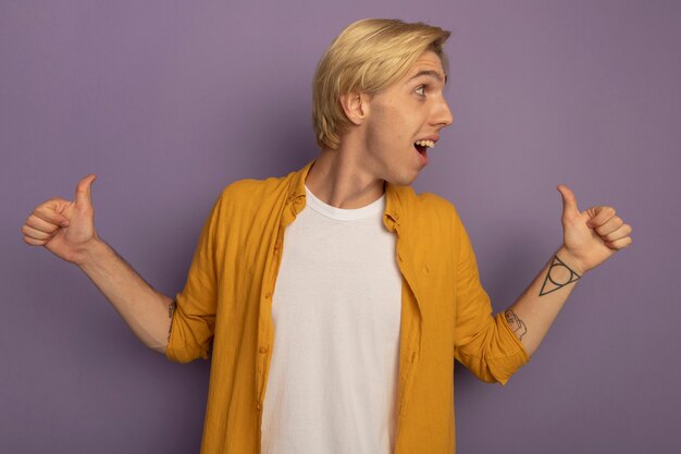 Smiling looking at side young blonde guy wearing yellow t-shirt points at himself isolated on purple