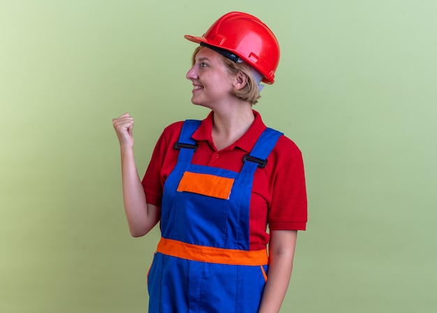 smiling looking side young builder woman in uniform showing yes gesture isolated on olive green wall