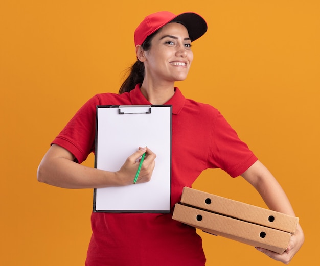 Sorridente guardando a lato la giovane ragazza delle consegne che indossa l'uniforme e il cappuccio che tiene gli appunti con scatole per pizza isolate sulla parete arancione