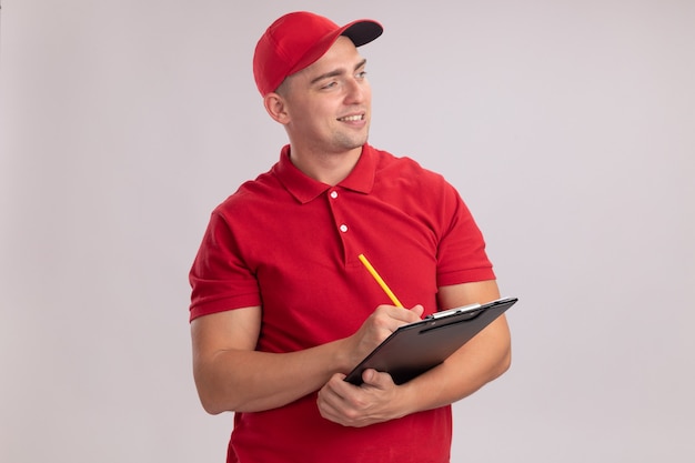 Smiling looking at side young delivery man wearing uniform with cap writing something in clipboard isolated on white wall