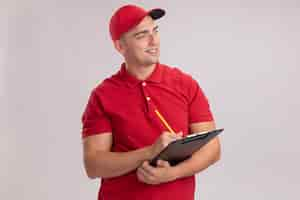 Foto gratuita sorridente guardando al lato giovane uomo di consegna indossando l'uniforme con cappuccio scrivendo qualcosa negli appunti isolato sul muro bianco