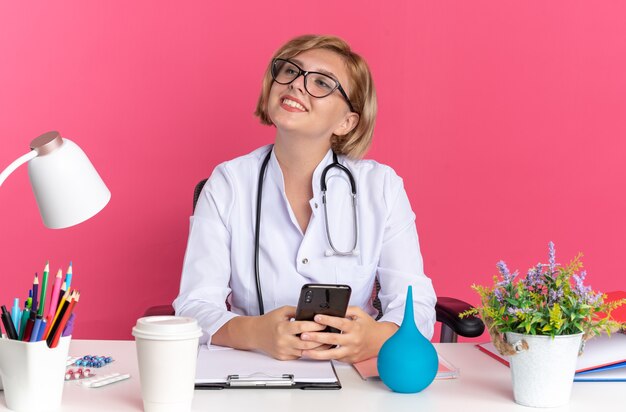 Smiling looking side young female doctor wearing medical robe with stethoscope and glasses sits at desk with medical tools holding phone isolated on pink background