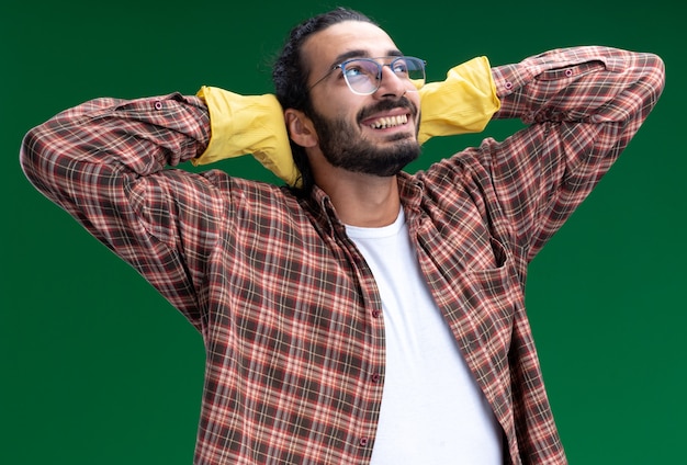 Smiling looking at side young handsome cleaning guy wearing t-shirt and gloves putting hands behind head isolated on green wall