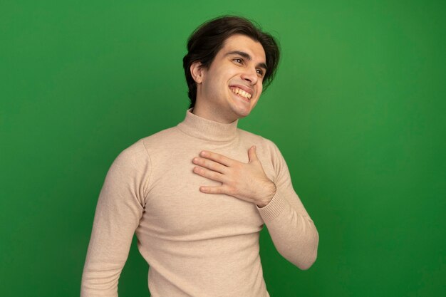 Smiling looking at side young handsome guy putting hand on chest isolated on green wall with copy space