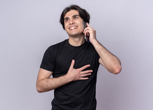 Smiling looking at side young handsome guy wearing black t-shirt speaks on phone isolated on white wall