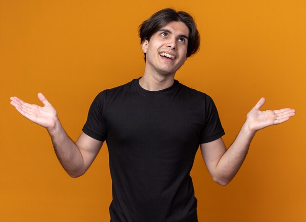 Smiling looking at side young handsome guy wearing black t-shirt spreading hands isolated on orange wall