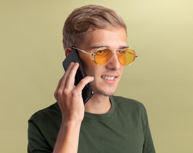 Улыбающийся, смотрящий в сторону молодой красивый парень в зеленой рубашке с очками говорит по телефону, изолированному на оливково-зеленой стене