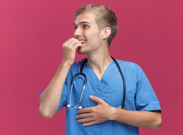 Smiling looking at side young male doctor wearing doctor uniform with stethoscope bites nails isolated on pink wall