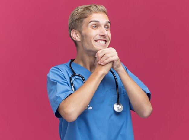 ピンクの壁に分離されたあごの下で手をつないで聴診器と医師の制服を着ている側の若い男性医師を見て笑顔