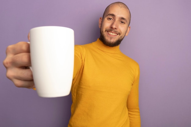 Sorridente guardando dritto giovane bel ragazzo che tiene tazza di tè isolato sulla porpora