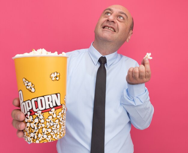 Smiling looking up middle-aged man wearing white t-shirt with tie holding bucket of popcorn with popcorn piece isolated on pink wall