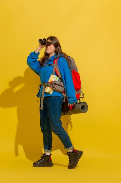 Smiling, looking up. Portrait of a cheerful young caucasian tourist girl with bag and binoculars isolated on yellow studio background. Preparing for traveling. Resort, human emotions, vacation.