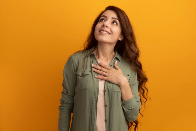 Smiling looking up young beautiful girl wearing olive green t-shirt putting hand on heart isolated on yellow wall