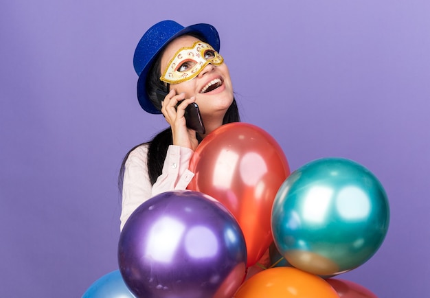 Smiling looking up young beautiful girl wearing party hat and masquerade eye mask standing behind balloons speaks on phone isolated on blue wall