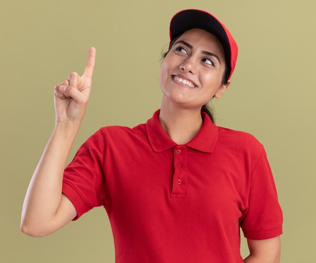 Smiling looking at up young delivery girl wearing uniform with cap points at up isolated on olive green wall