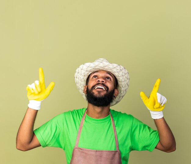 Smiling looking up young gardener afro-american guy wearing gardening hat with gloves points at up isolated on olive green wall