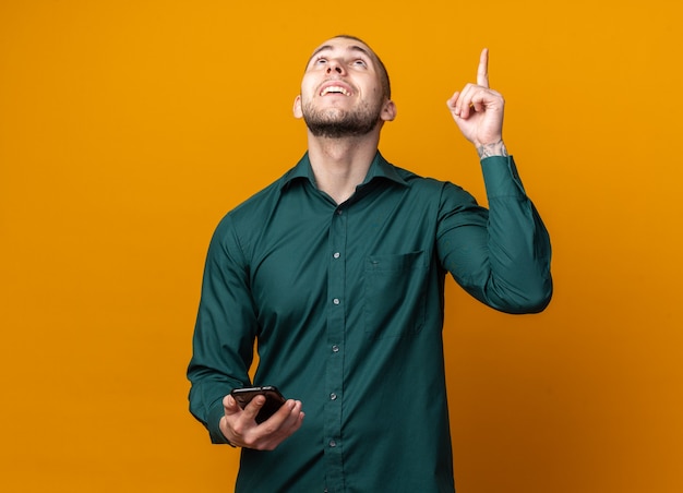 Smiling looking up young handsome guy wearing green shirt holding phone points at up 