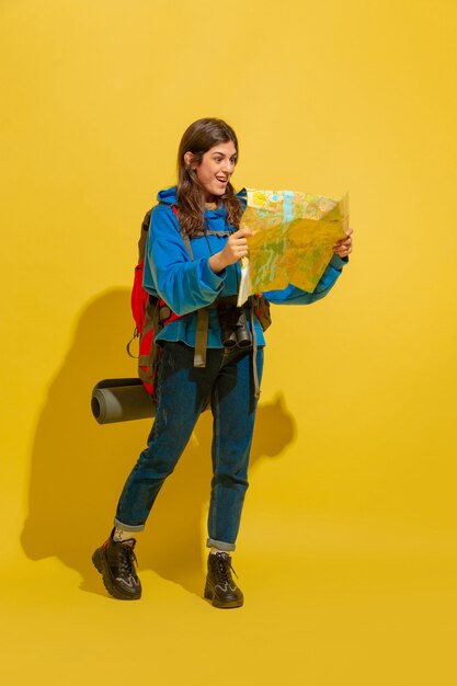 Smiling, looking for way. Portrait of a cheerful young caucasian tourist girl with bag and binoculars isolated on yellow studio background. Preparing for traveling. Resort, human emotions, vacation.