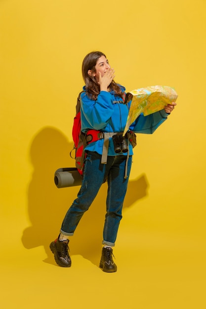 Улыбается, ищет путь. Портрет веселой молодой кавказской туристической девушки с сумкой и биноклем, изолированными на желтом фоне студии.