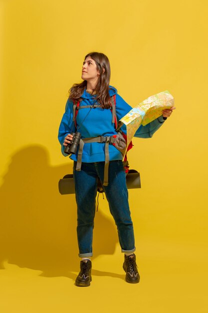 Smiling, looking for way. Portrait of a cheerful young caucasian tourist girl with bag and binoculars isolated on yellow studio background.