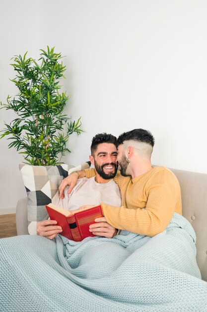 Smiling loving young gay couple lying on sofa in one blue blanket