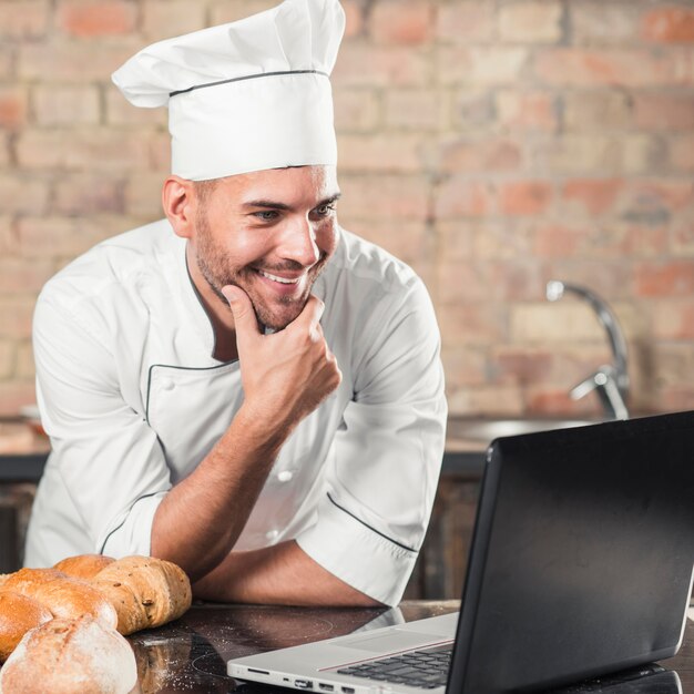 Smiling male baker leaning on kitchen worktop looking at laptop