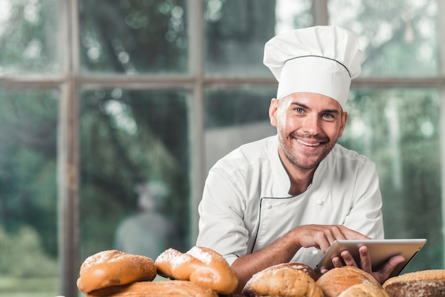 Улыбаясь мужской пекарь, опираясь на стол с испеченным хлебом против окна