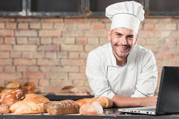 Free photo smiling male baker with different type of baked breads and laptop on kitchen worktop