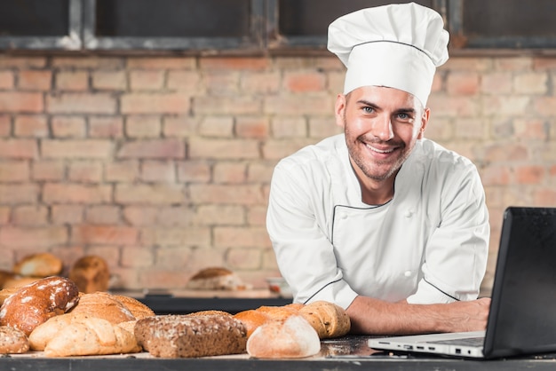 Smiling male baker with different type of baked breads and laptop on kitchen worktop