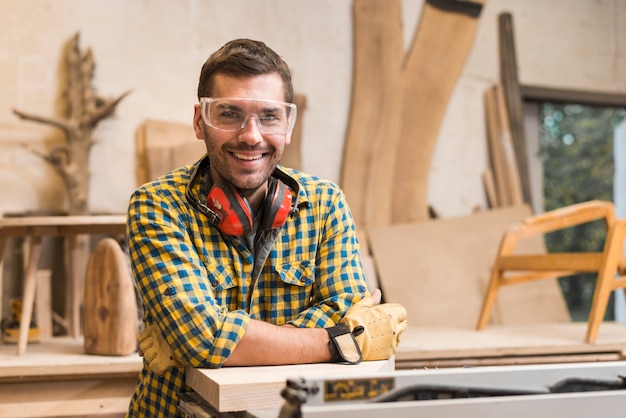 Улыбающийся мужчина-плотник с ушным защитником на шее стоит в своей мастерской