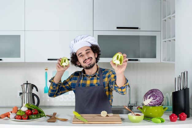 新鮮な野菜で男性シェフを笑顔にし、キッチンツールで調理し、白いキッチンでカットピーマンを保持します
