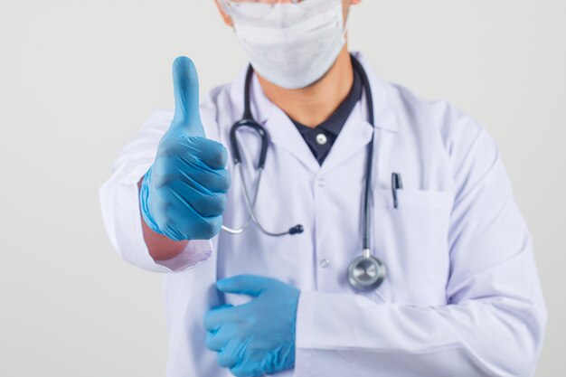 Smiling male doctor in white coat and mask making thumb up and looking happy