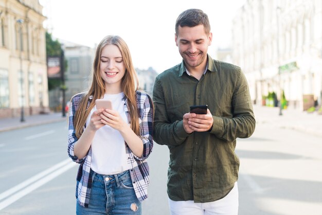 Улыбающиеся друзья мужского и женского пола, с помощью мобильного телефона белый ходить вместе на улице