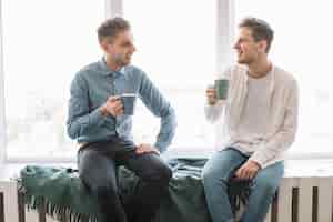 Бесплатное фото Улыбающиеся друзья-мужчины, сидящие у окна, пьющие кофе