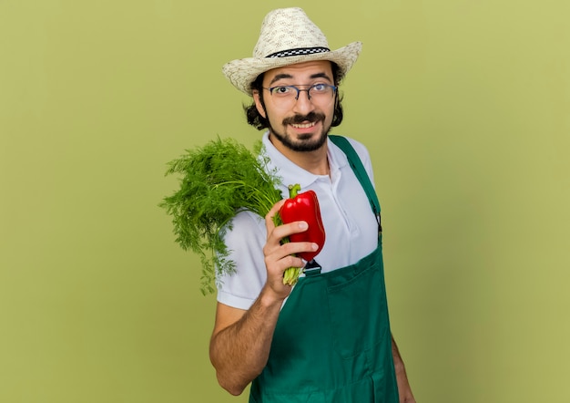 Smiling male gardener in optical glasses wearing gardening hat holds fennel and red pepper