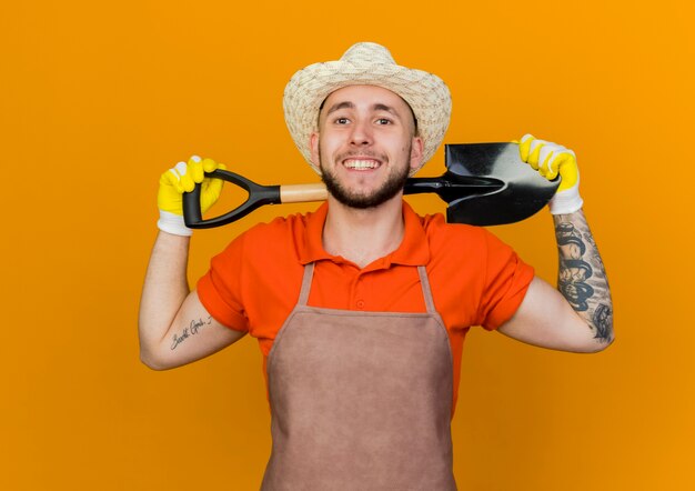 Smiling male gardener wearing gardening hat and gloves holds spade behind neck looking  