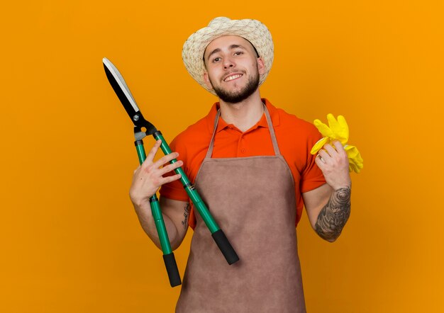 Smiling male gardener wearing gardening hat holds clippers and gloves 