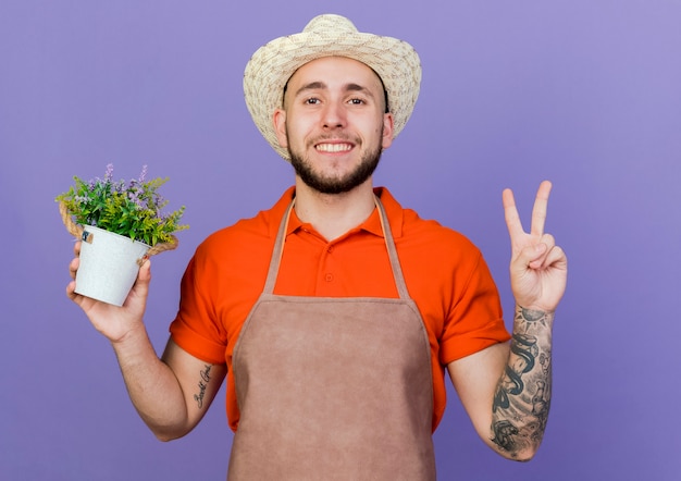 Sorridente giardiniere maschio che indossa un cappello da giardinaggio tiene un vaso di fiori e fa un gesto con il segno della mano della vittoria