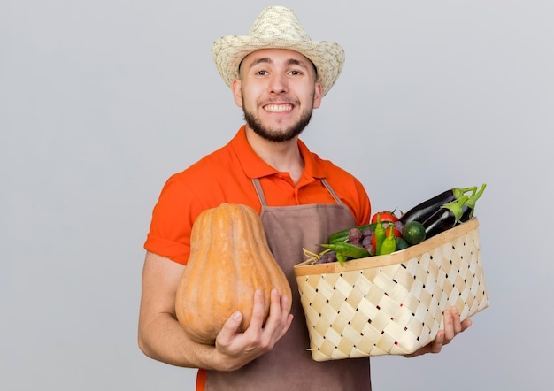 Smiling male gardener wearing gardening hat holds pumpkin and vegetable basket looking  