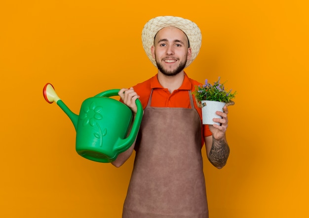 Smiling male gardener wearing gardening hat holds watering can and flowers in flowerpot 