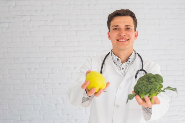 Smiling male happy doctor offering apple and broccoli