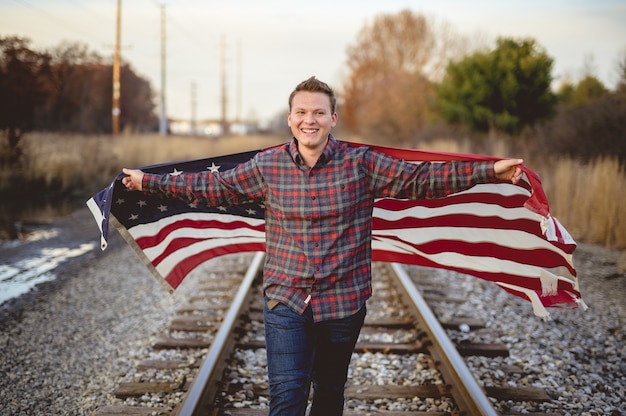 Free photo smiling male holding the united states flag while walking on the train rails