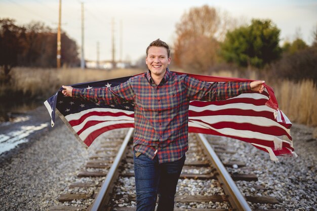 Улыбающийся мужчина держит флаг США во время прогулки по рельсам поезда