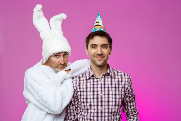 Smiling male and man in rabbit costume at birthday party over purple wall.