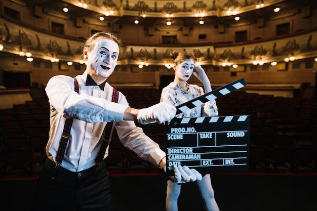 Free photo smiling male mime artist holding clapperboard in front of female mime on stage