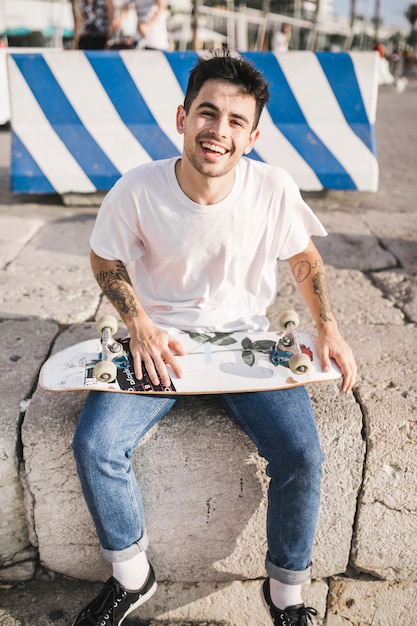 Foto gratuita skateboarder maschio sorridente che si siede con il pattino davanti a barricata