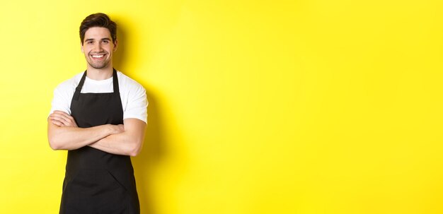 Smiling male waiter in black apron standing confident cross arms on chest against yellow background