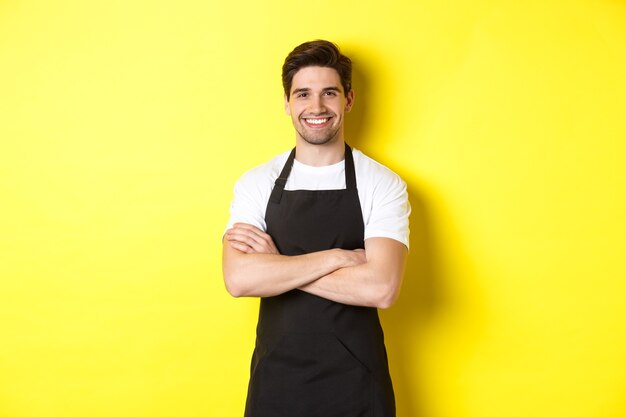 Smiling male waiter in black apron standing confident cross arms on chest against yellow background