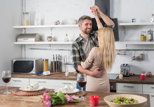 Smiling man dancing with blond woman near table in kitchen